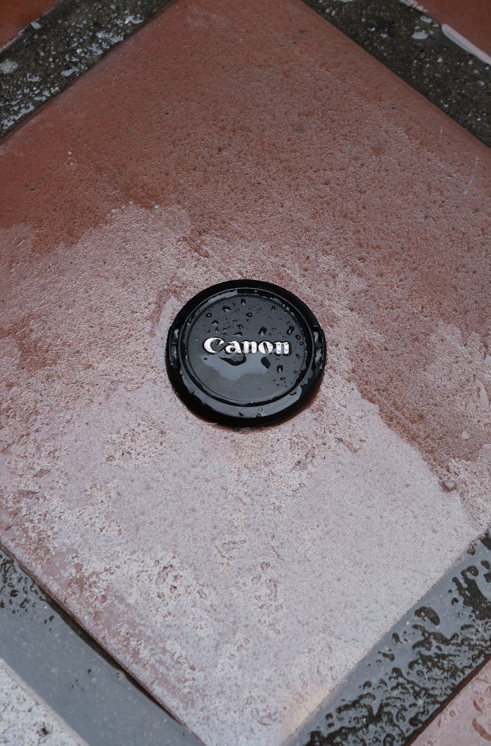Close-up of a Canon lens cap covered in raindrops on a wet square tile surface.