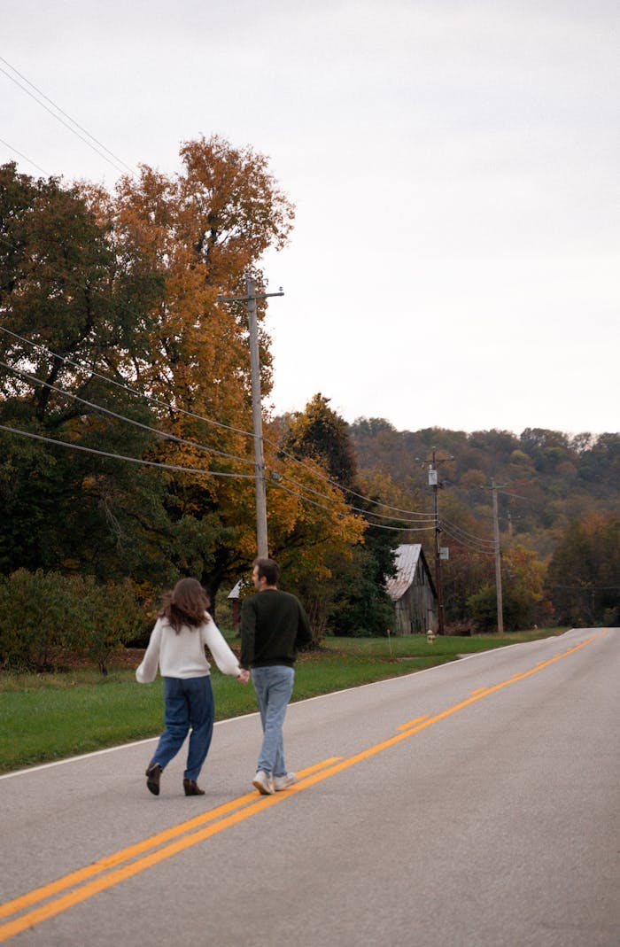 A couple holds hands while walking on a scenic road amidst vibrant fall foliage in Kentucky.